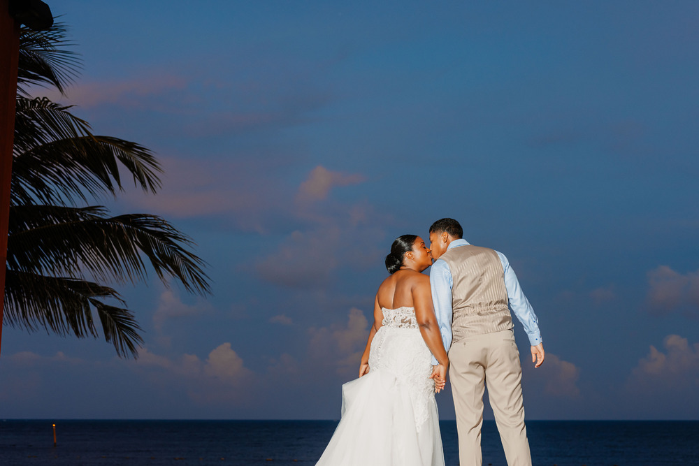 Jessie and Taylor share a kiss in wedding attire by the ocean at sunset during their destination wedding.