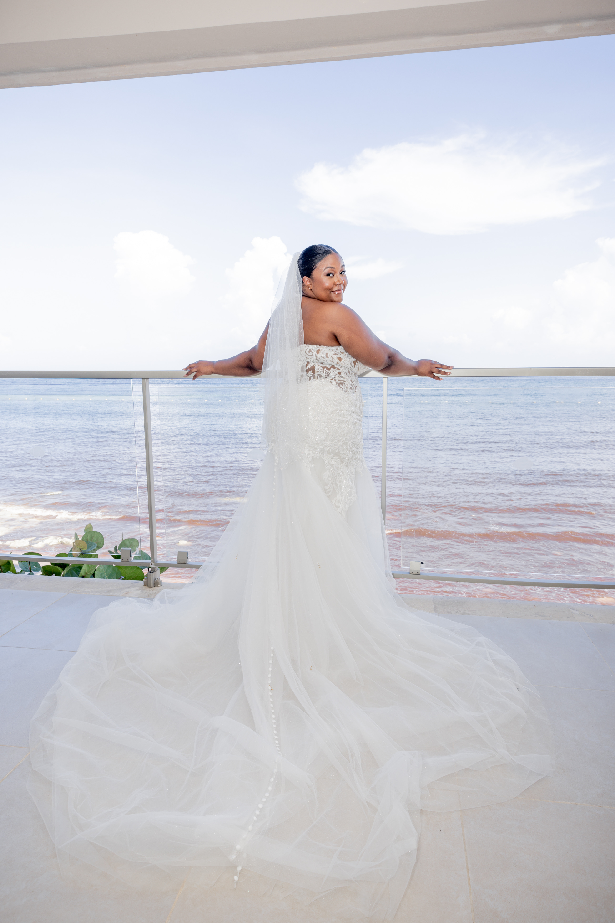 Bride in April and Javiert wedding gown stands on balcony at a sunny destination wedding by the ocean.