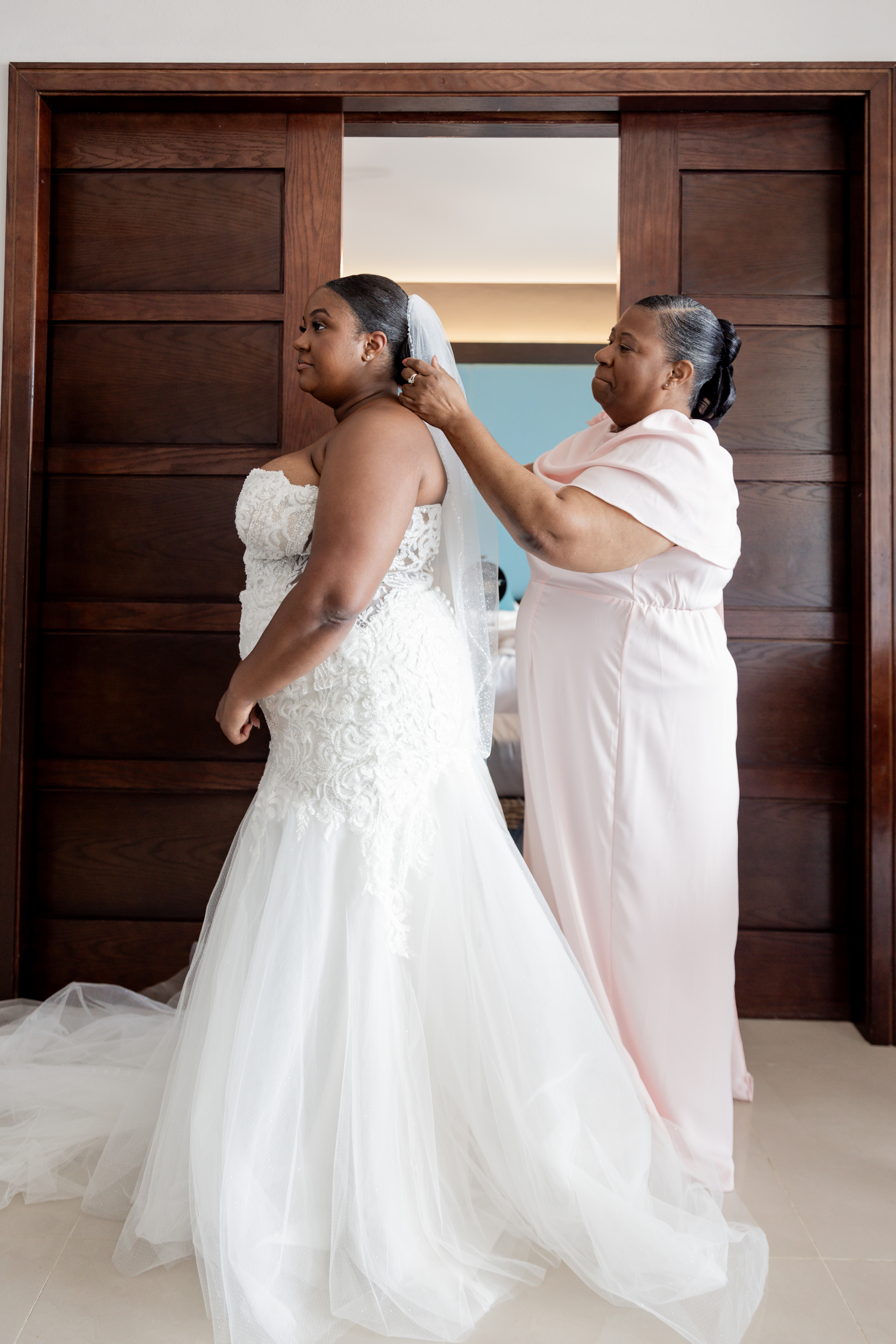 A woman helps a bride with her April and Javiert veil before a destination wedding, near wooden doors.