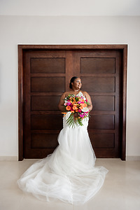 Bride in April and Javiert gown holds vibrant bouquet before wooden doors at a destination wedding.