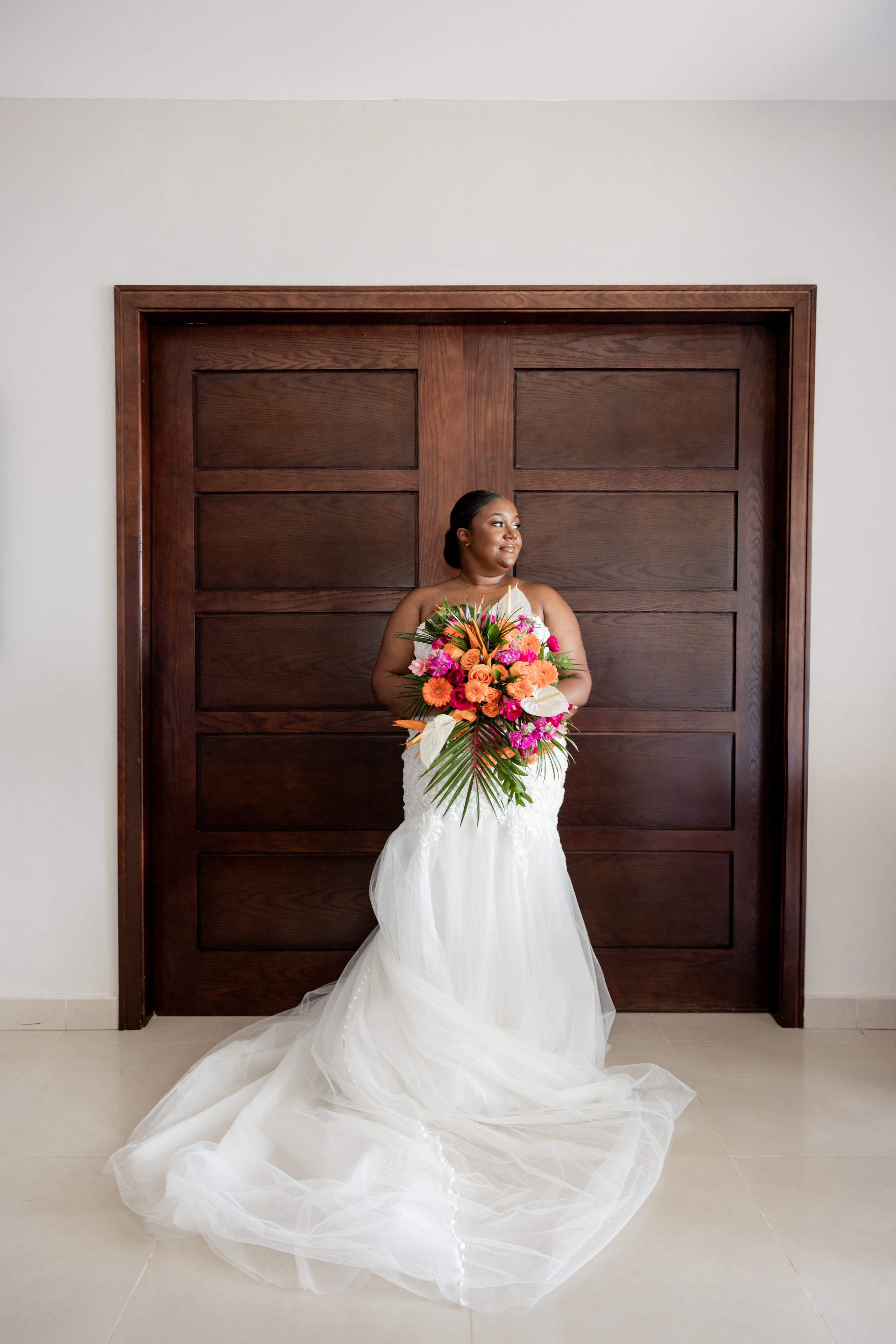 Bride in April and Javiert gown holds vibrant bouquet before wooden doors at a destination wedding.