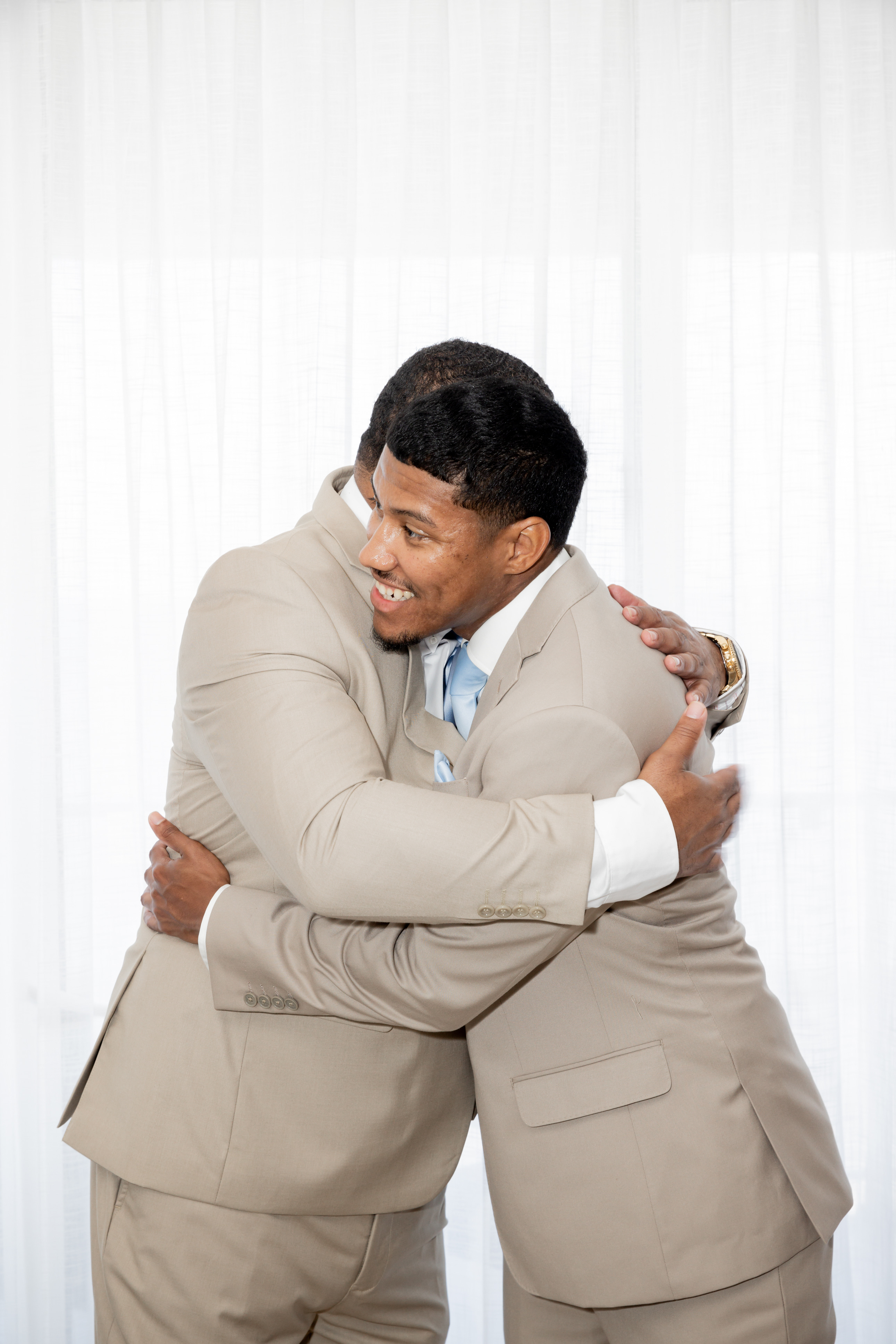 April and Javiert, in beige suits, embrace at a destination wedding in front of a white curtain.