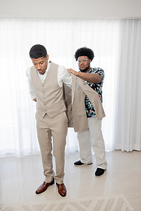 In a bright room, a man assists another in putting on an April and Javiert beige suit for a wedding.
