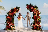 Jessie and Taylor hold hands beneath a floral arch by the ocean at their sunny destination wedding.