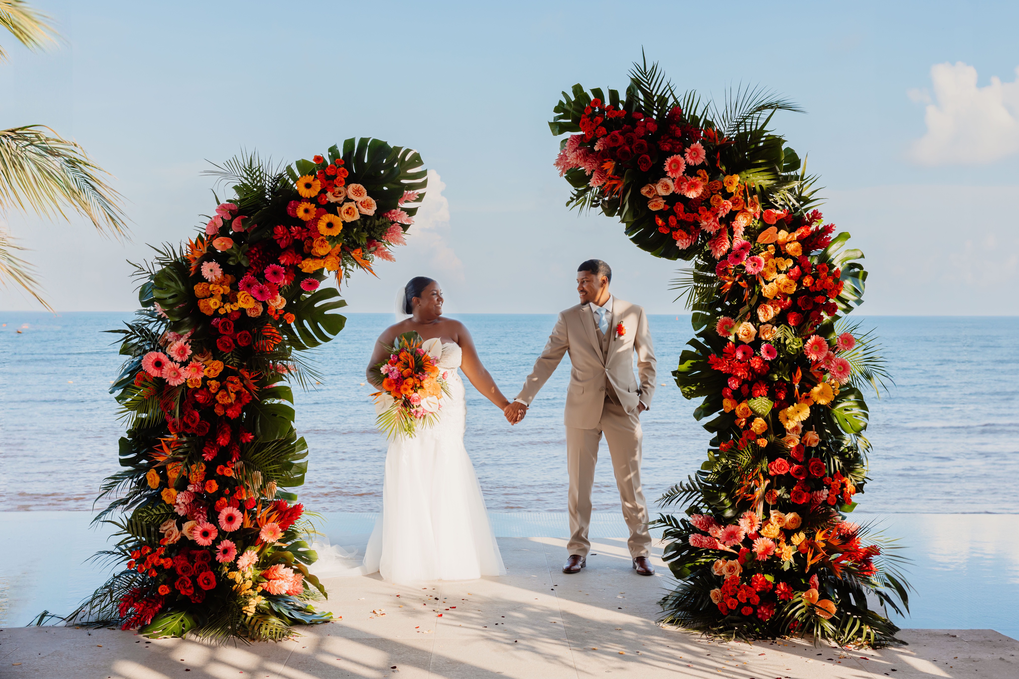 Jessie and Taylor hold hands beneath a floral arch by the ocean at their sunny destination wedding.