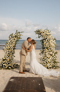 April and Javiert share a kiss at their destination wedding on the beach, framed by floral arches.