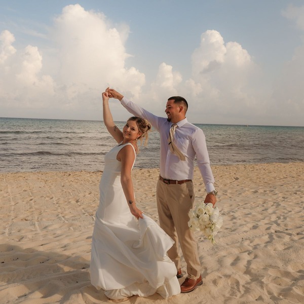 April and Javiert pose together on a sandy beach at their destination wedding with the ocean behind them.