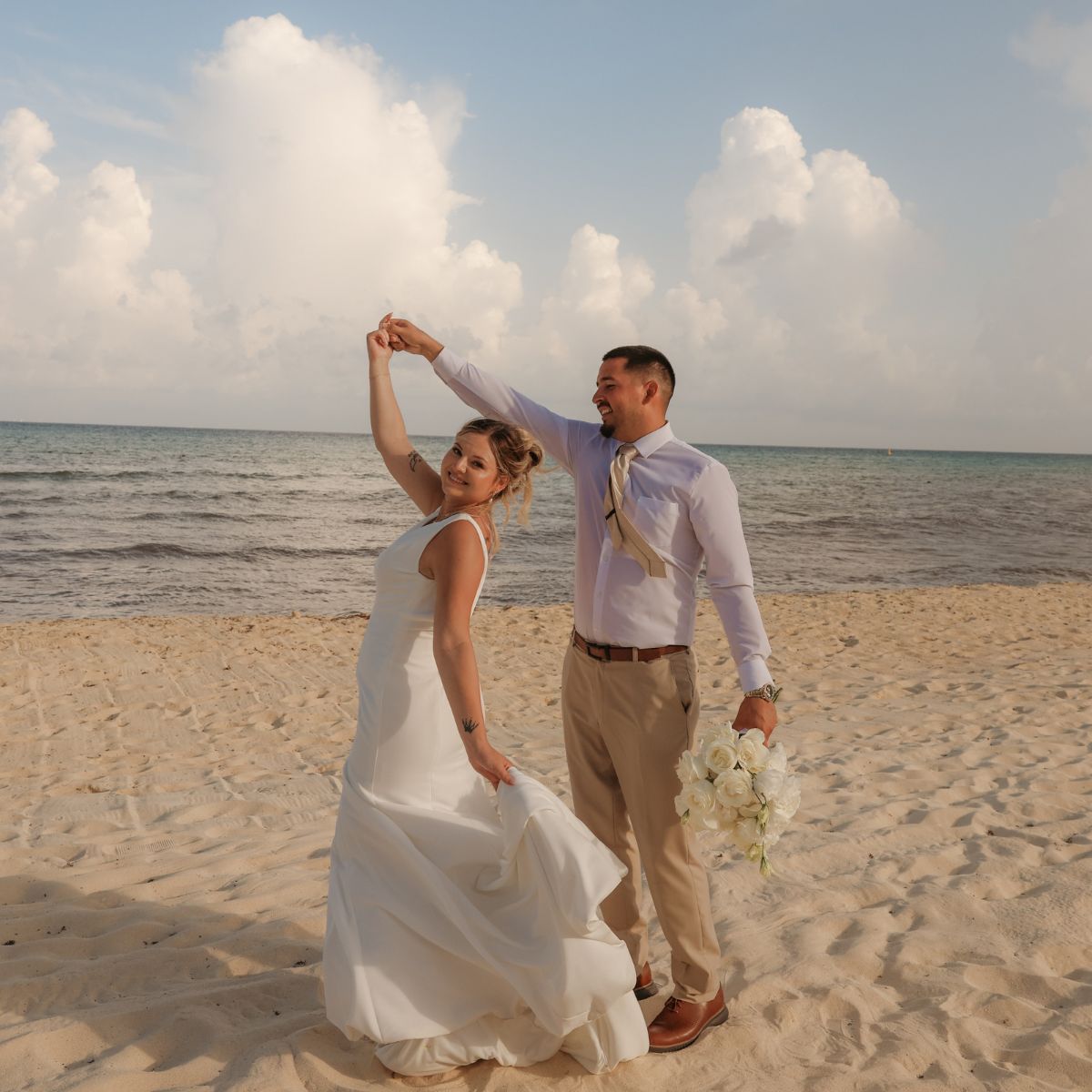 April and Javiert pose together on a sandy beach at their destination wedding with the ocean behind them.