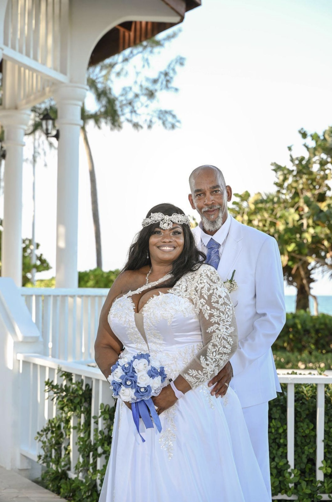 Bride and groom pose outdoors in wedding attire, holding a blue and white bouquet at their wedding.