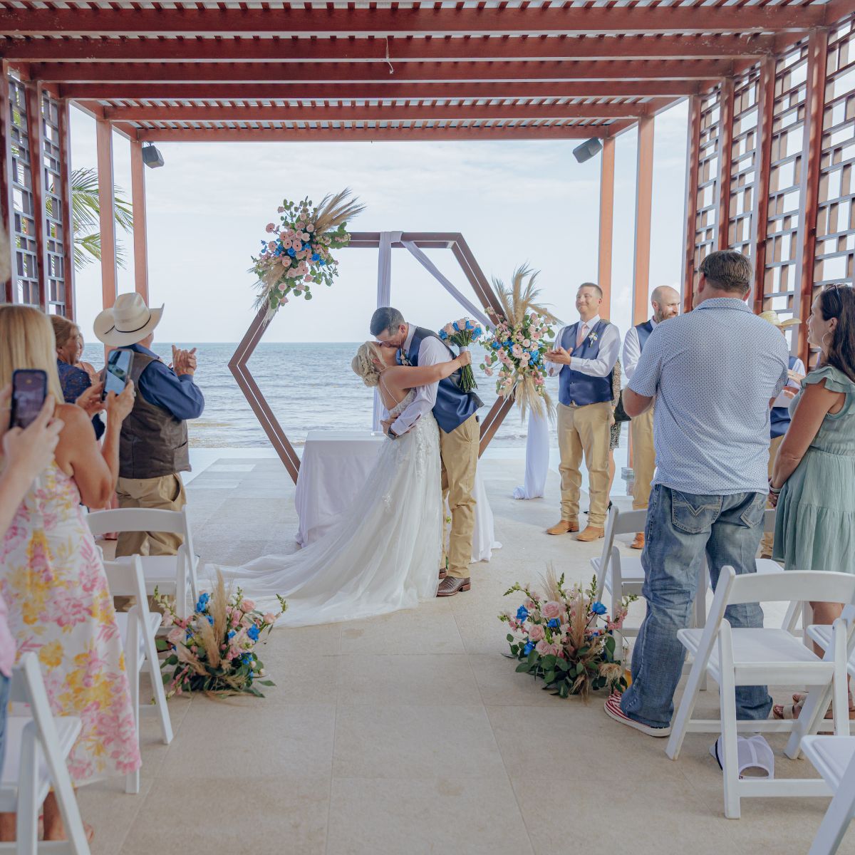 At a destination wedding, the bride and groom kiss outdoors while guests applaud their union.