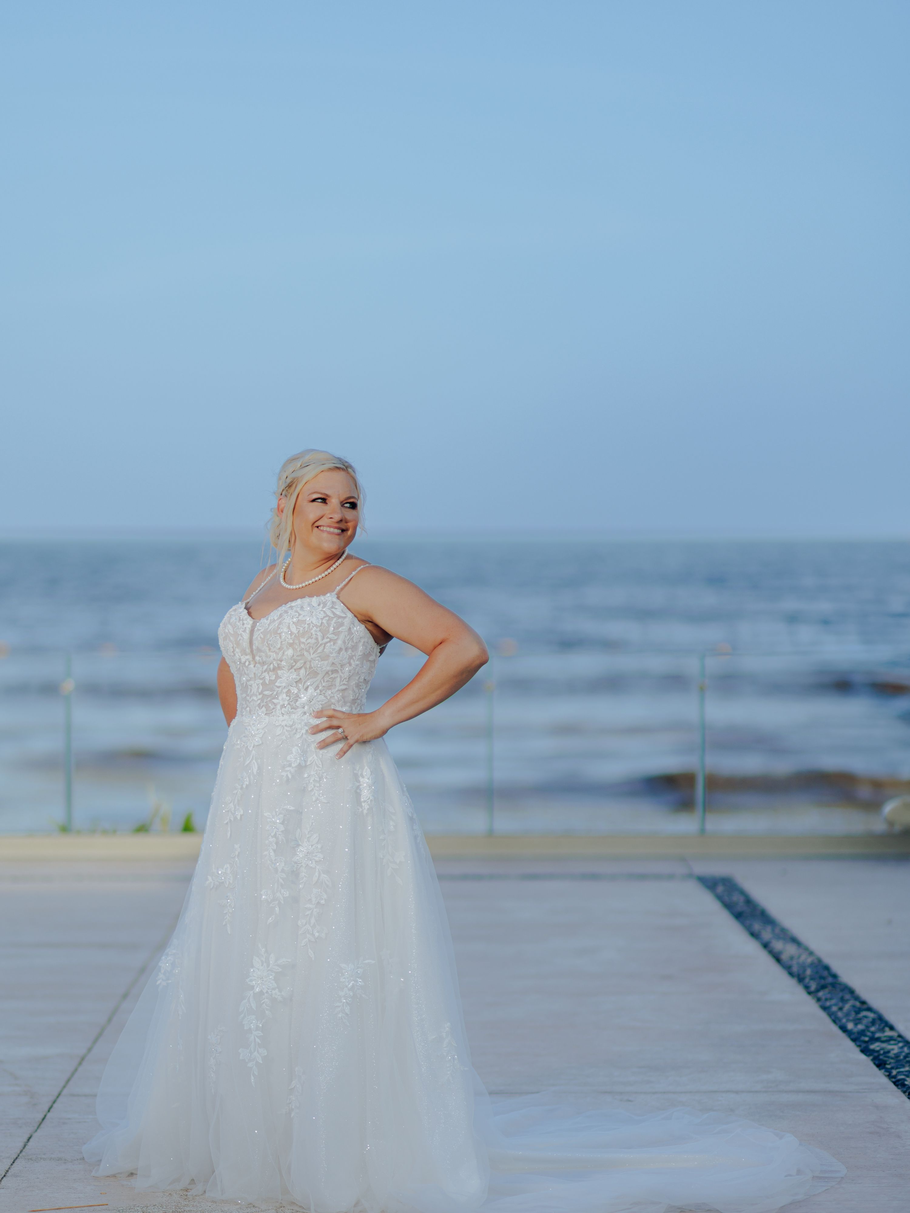 Bride in a white wedding dress poses and smiles by the ocean at a beautiful destination wedding.
