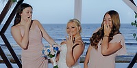 Three women in dresses smile by the ocean at a destination wedding, showing rings, one with a bouquet.