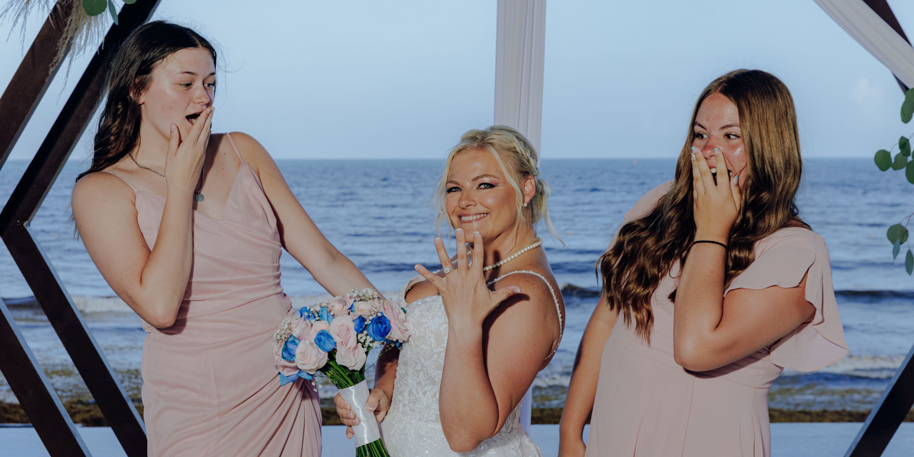 Three women in dresses smile by the ocean at a destination wedding, showing rings, one with a bouquet.
