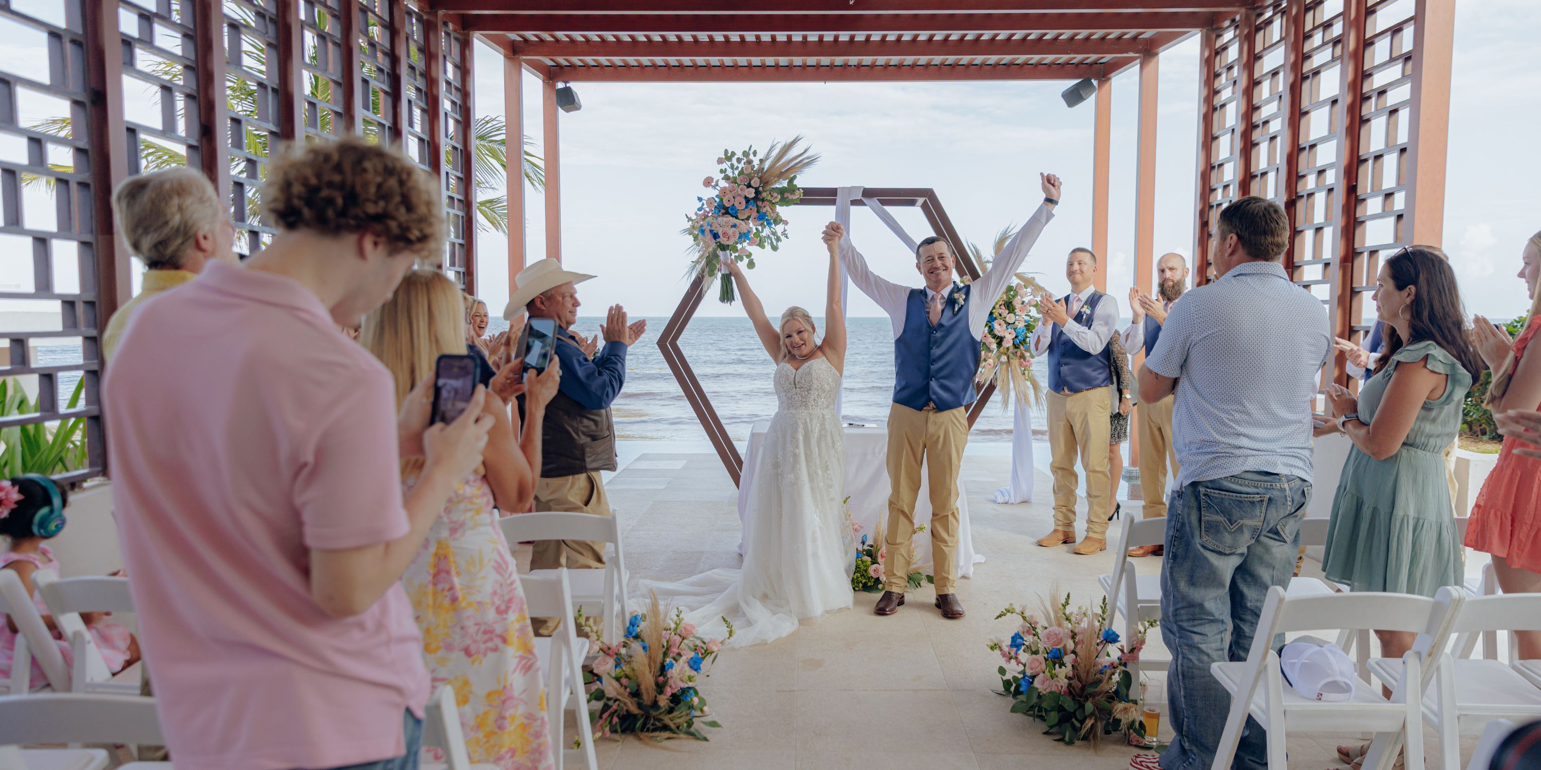 Bride and groom celebrate at the altar during a destination wedding as guests applaud outdoors.