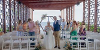 Bride and groom walk down the aisle at a destination wedding under a wooden pergola by the sea, guests watching.