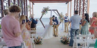 At a destination wedding by the ocean, newlyweds share a kiss while guests snap photos.