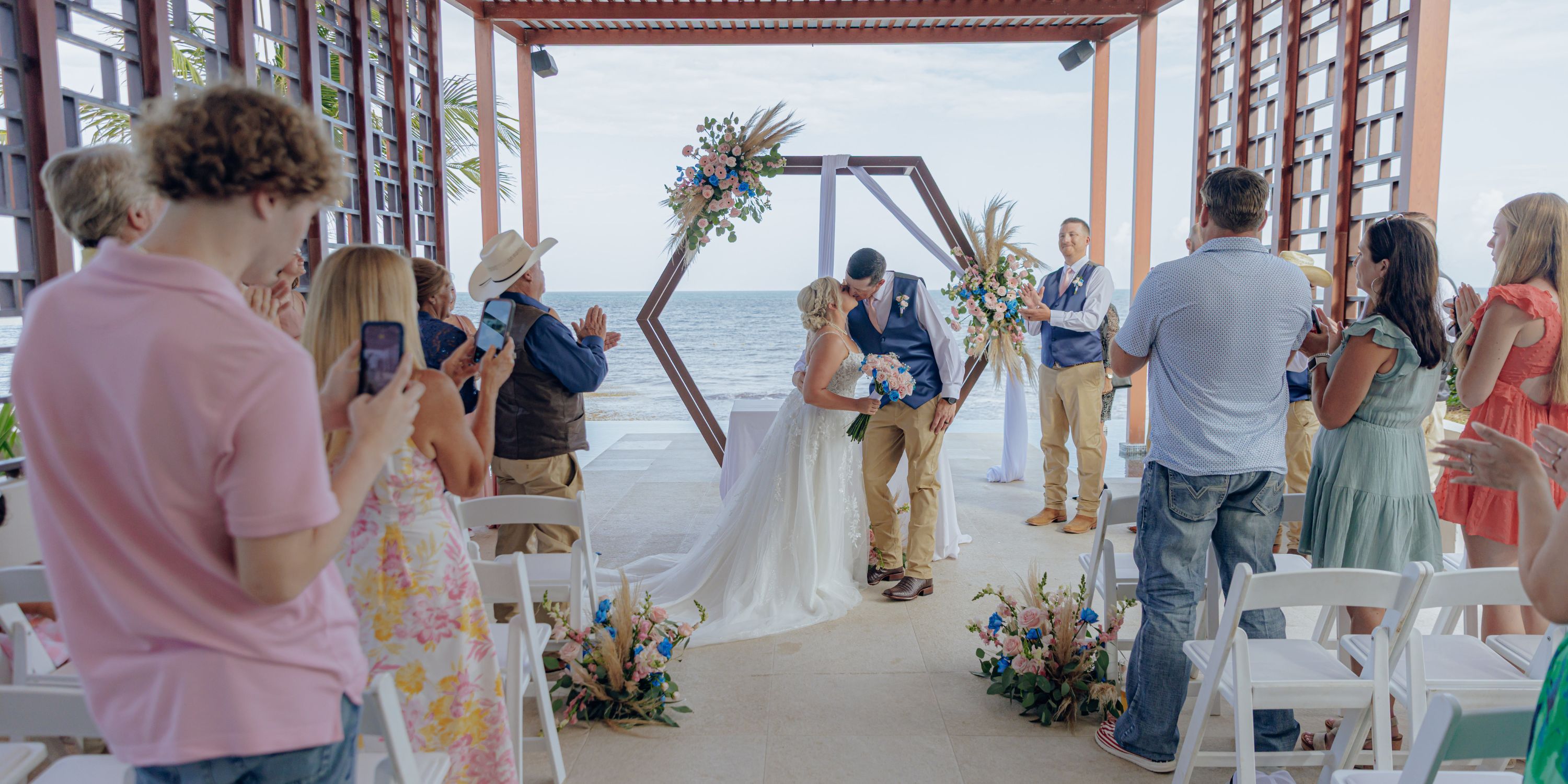 At a destination wedding by the ocean, newlyweds share a kiss while guests snap photos.