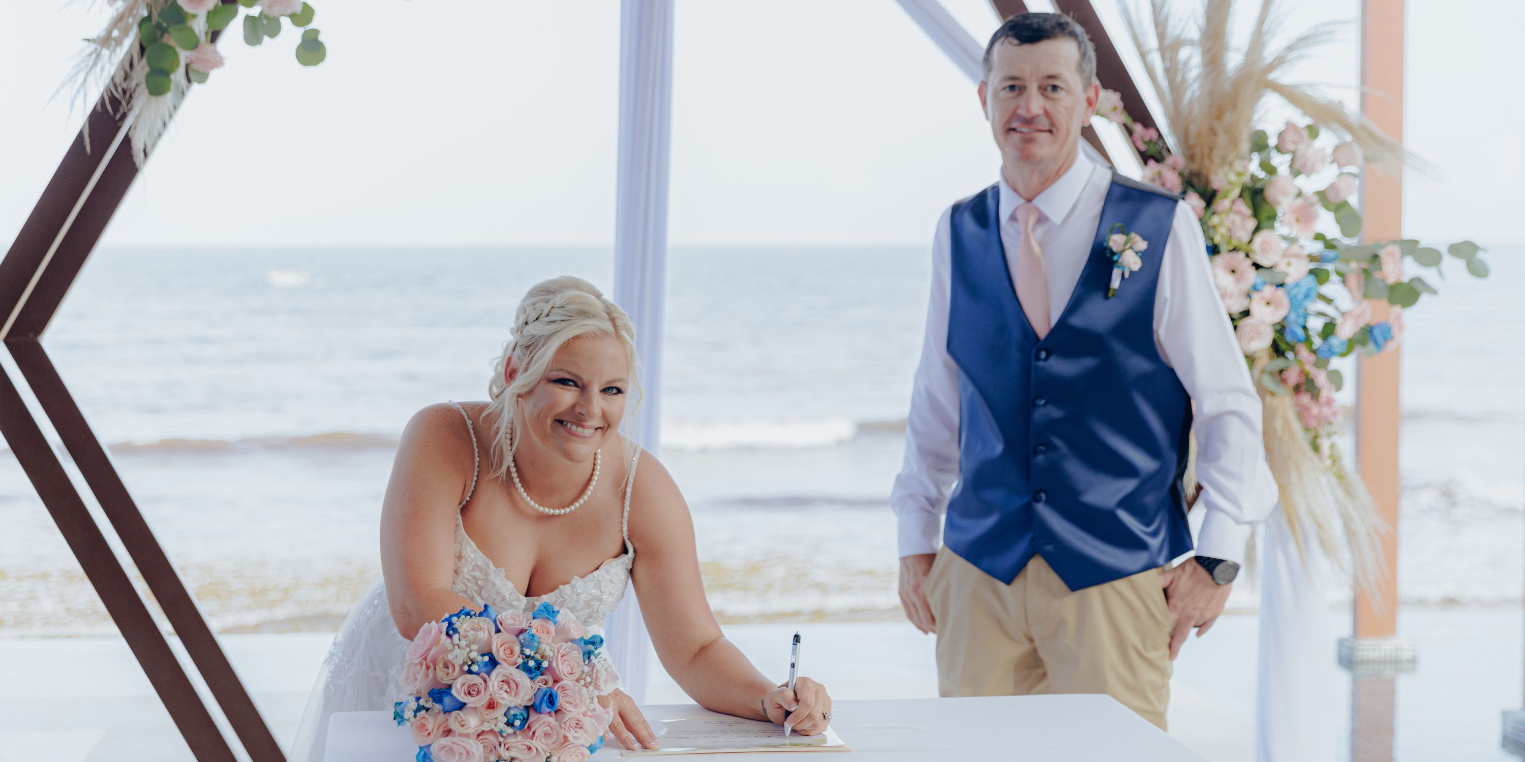 Bride signing a document at a destination wedding ceremony by the beach, groom standing next to her.