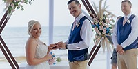 A destination wedding couple exchanges rings by the beach, framed by a floral backdrop and ocean views.