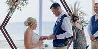 Bride and groom exchange rings at a destination wedding on the beach beneath a floral arch.