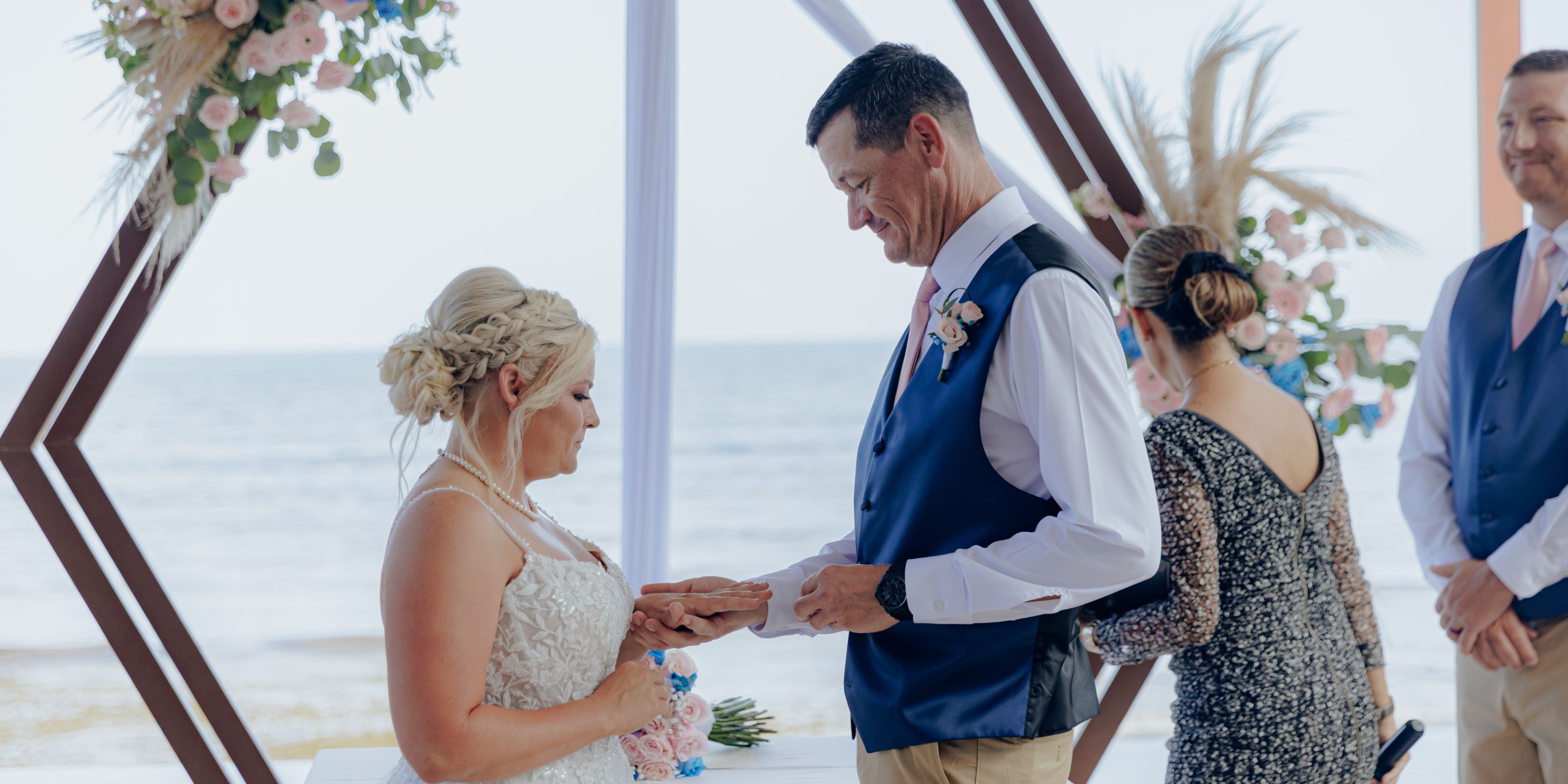Bride and groom exchange rings at a destination wedding on the beach beneath a floral arch.
