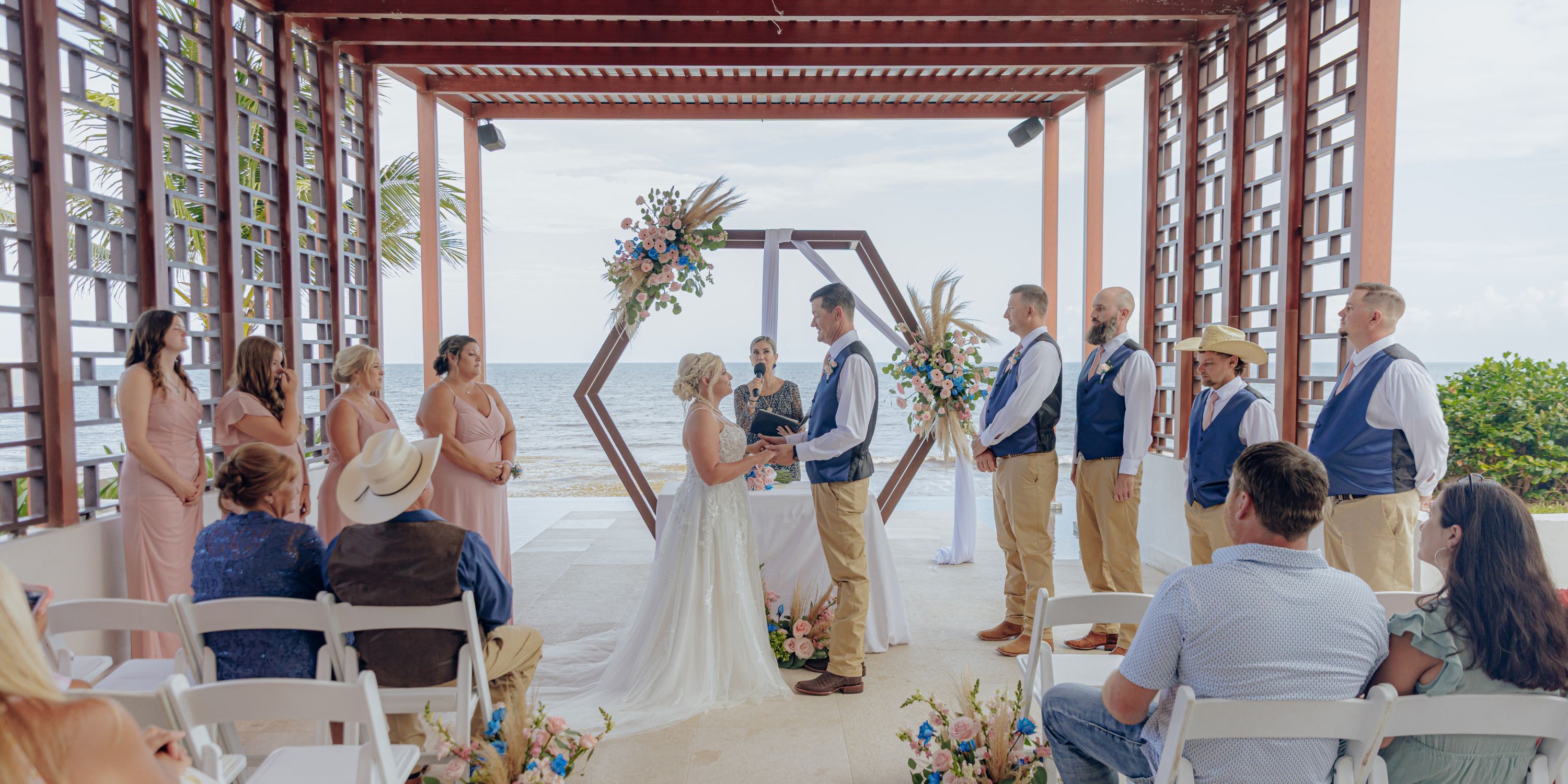 A couple exchange vows at their destination wedding, held outdoors with the ocean as a scenic backdrop.