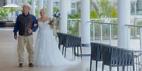 At a destination wedding, a bride in a white gown walks indoors beside an older man in casual western attire.