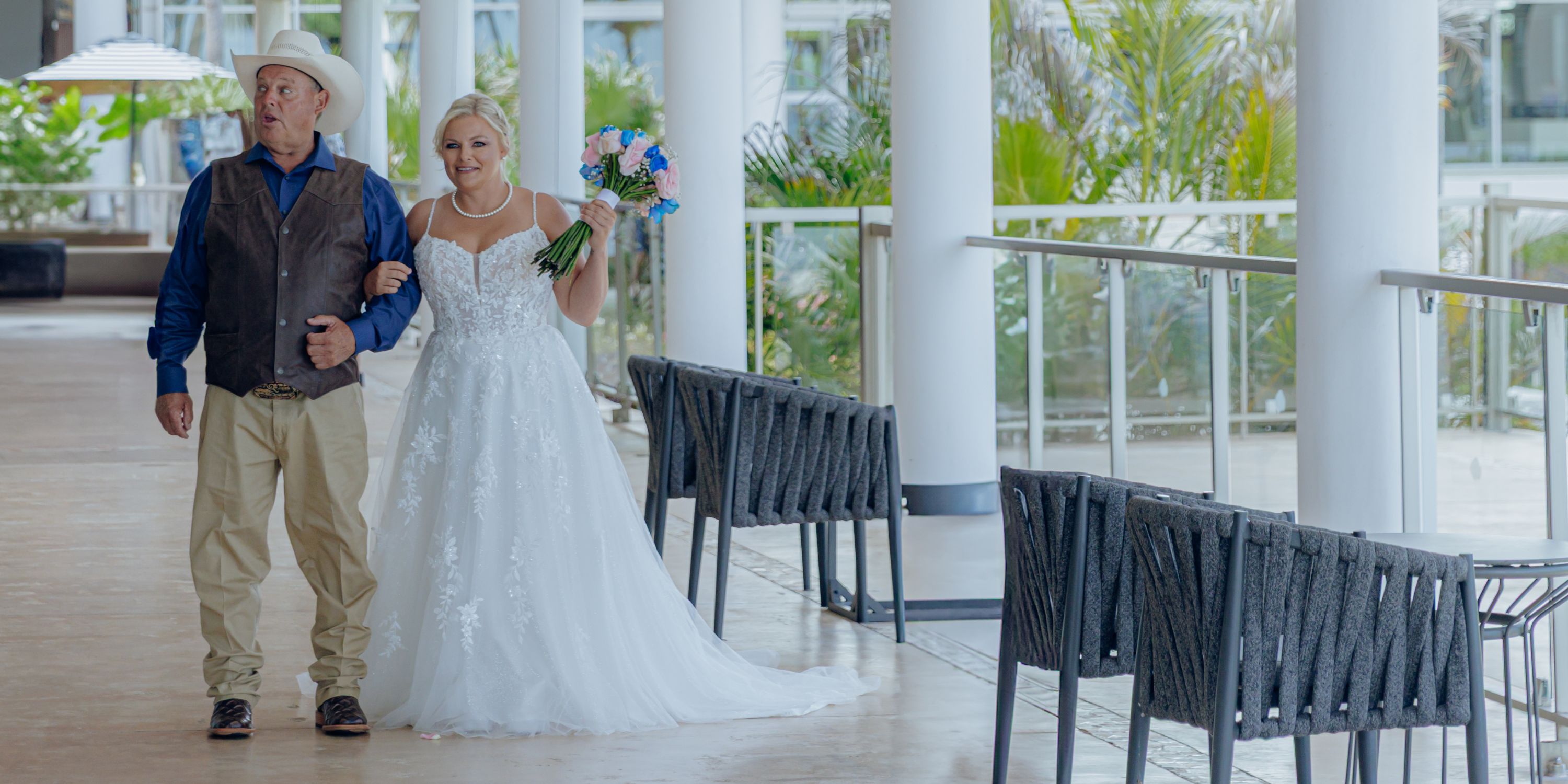 At a destination wedding, a bride in a white gown walks indoors beside an older man in casual western attire.