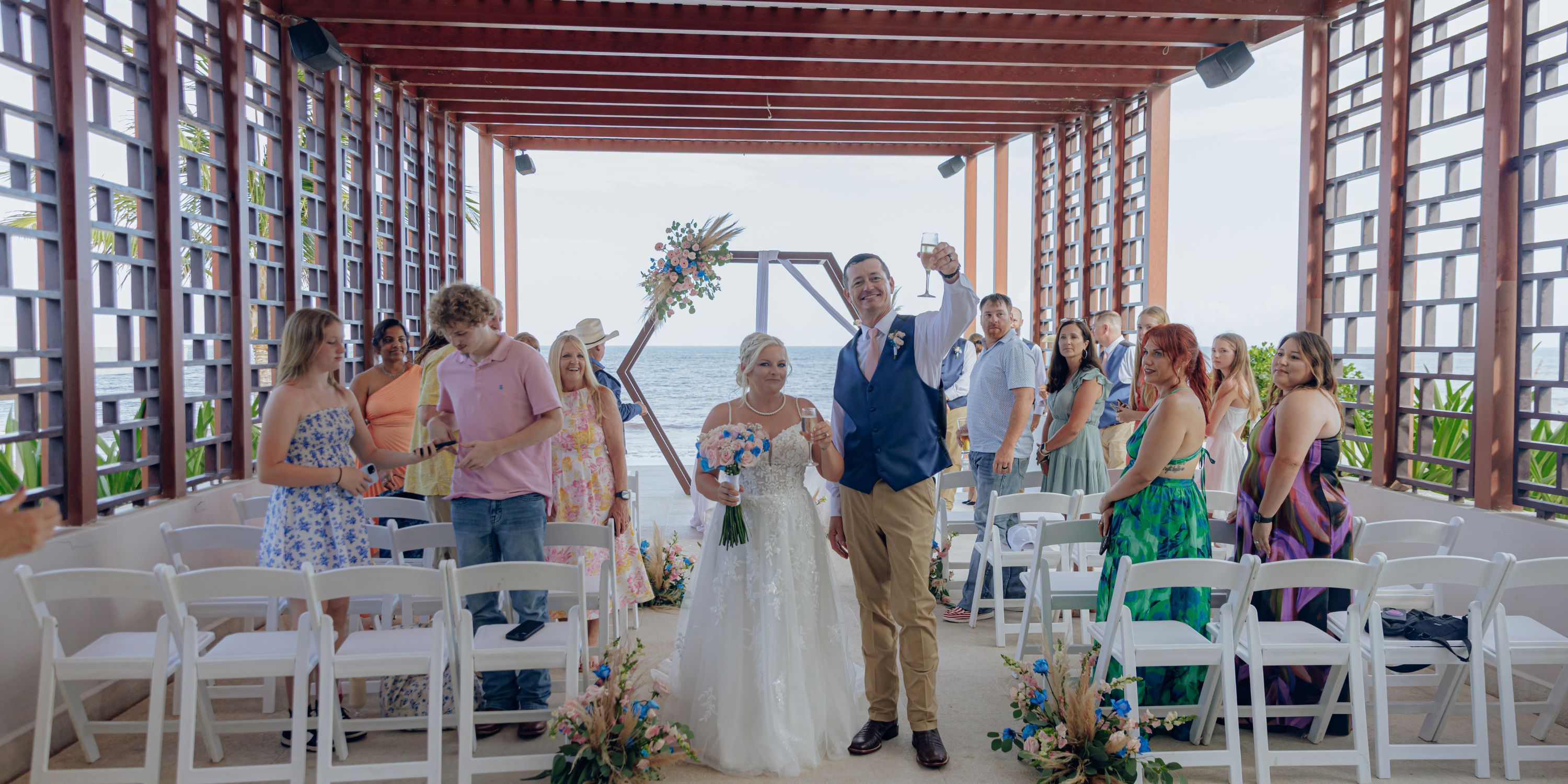 Bride and groom walk down the aisle at a destination wedding under a wooden pergola by the sea, guests watching.