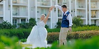 Bride and groom share a dance at their destination wedding, outdoors by a building amid lush greenery.