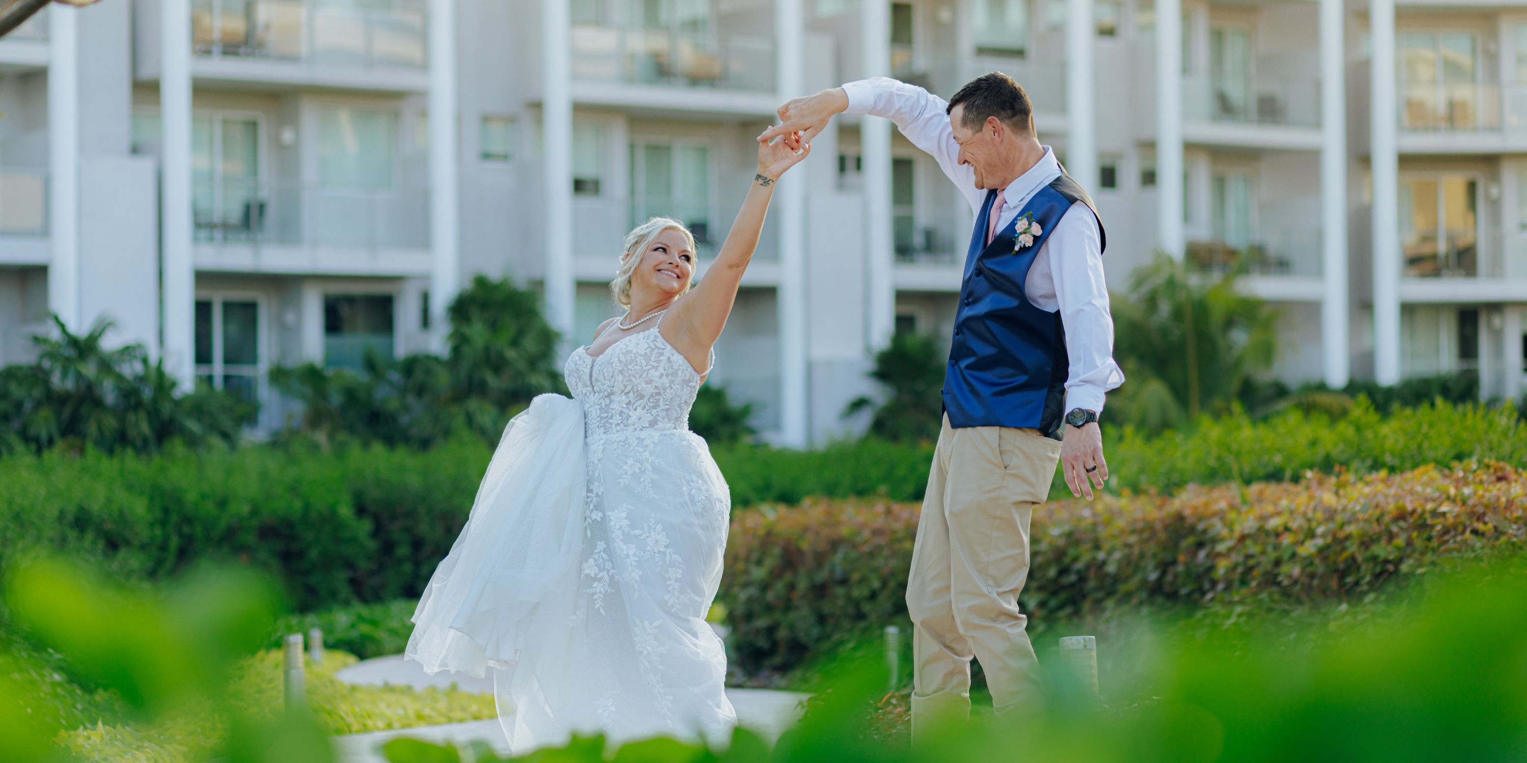 Bride and groom share a dance at their destination wedding, outdoors by a building amid lush greenery.
