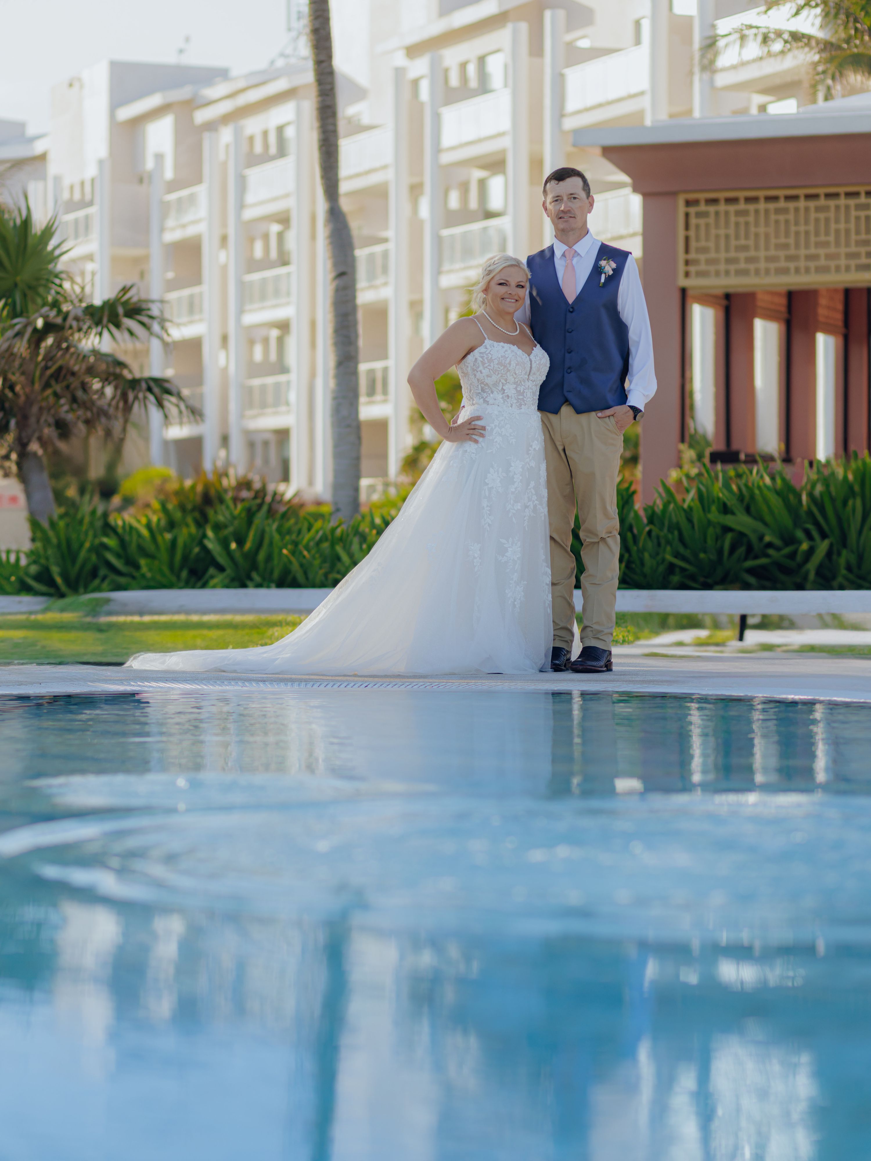 Bride and groom at their destination wedding, standing by a pool with a building behind them.