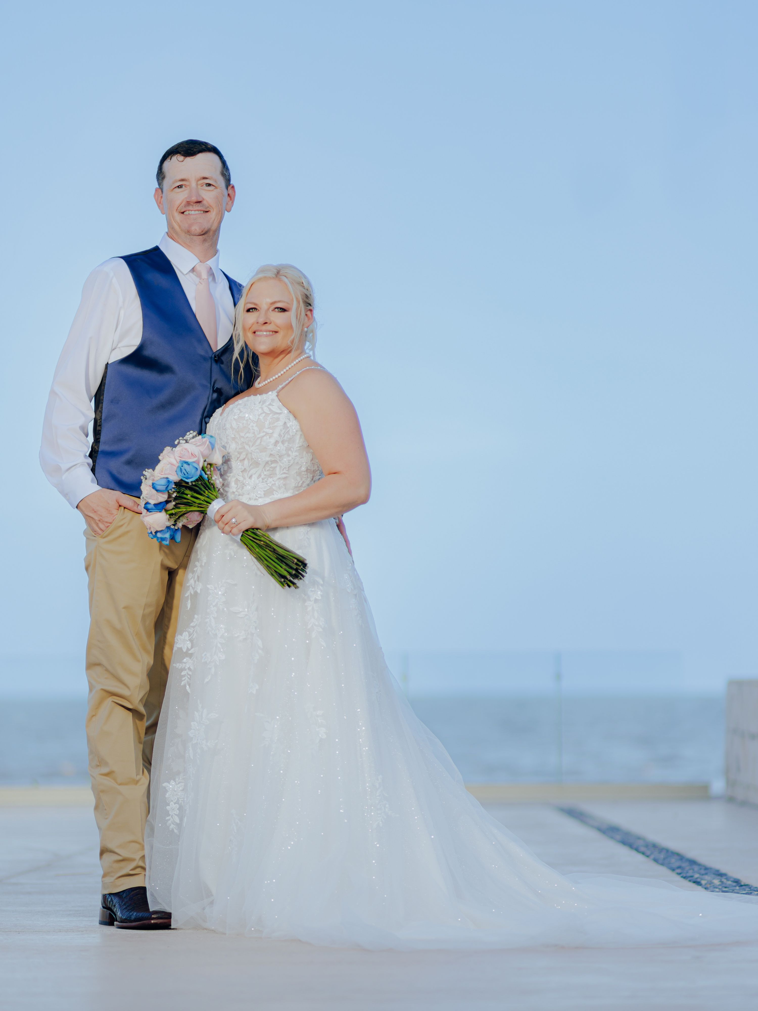 Bride and groom at their destination wedding stand outdoors holding flowers, ocean visible behind them.