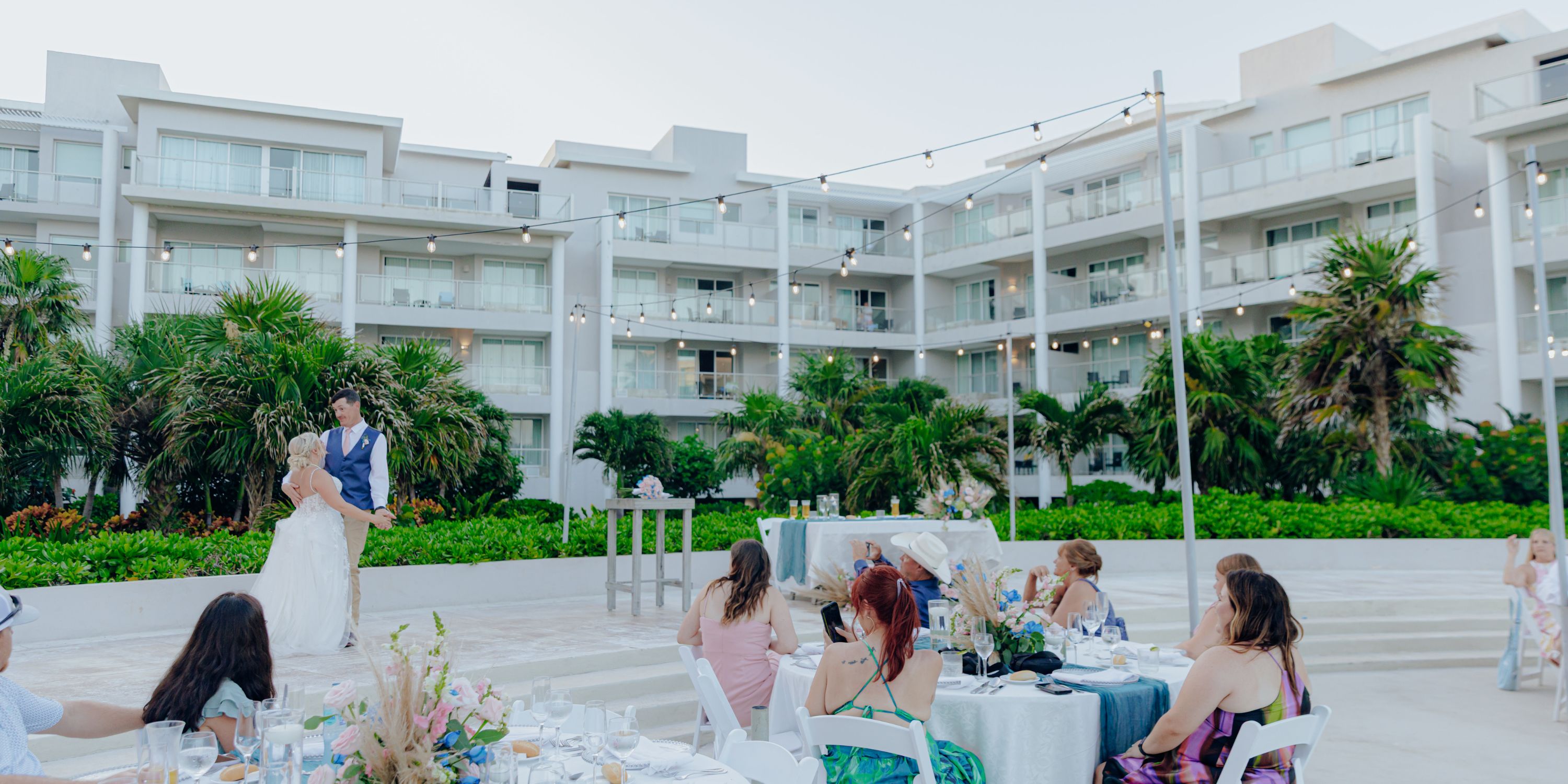 A couple dances at their destination wedding outdoors, surrounded by seated guests under string lights.