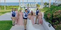 A destination wedding party poses outdoors by the ocean, with a "DREAMS" sign in the background.