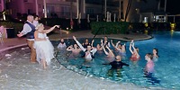 At a destination wedding, the bride tosses her bouquet to guests in and around the hotel pool at night.