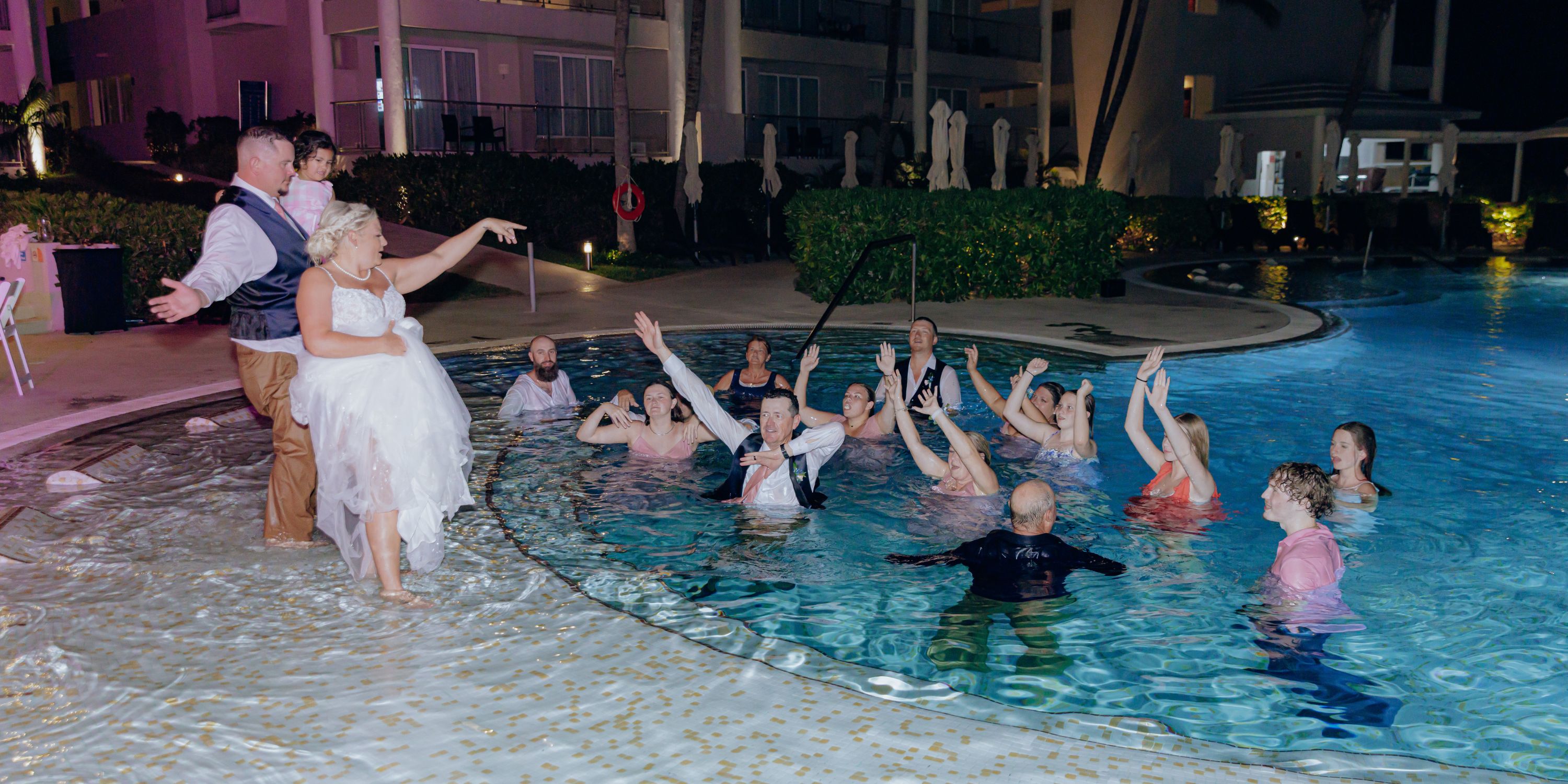 At a destination wedding, the bride tosses her bouquet to guests in and around the hotel pool at night.