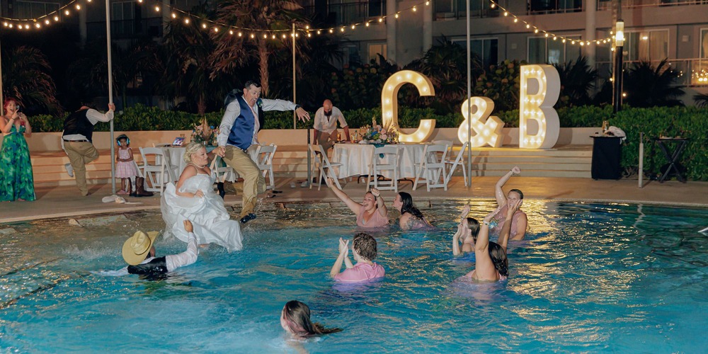 At a destination wedding, the bride, groom, and guests joyfully jump into a pool during the reception.