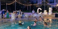 Newlyweds jump into a pool at their destination wedding at night; glowing C & B letters behind them.