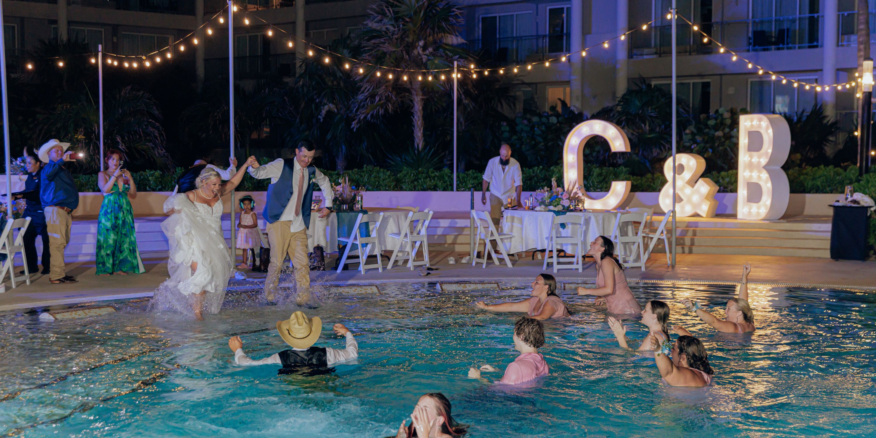 Newlyweds jump into a pool at their destination wedding at night; glowing C & B letters behind them.