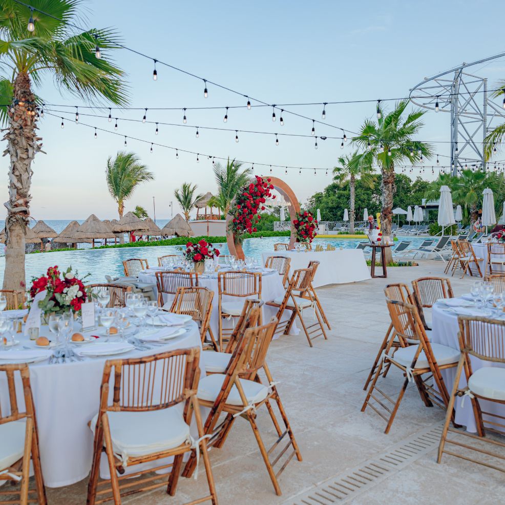 Elegant destination wedding reception by a pool, featuring round tables, chairs, and floral accents.