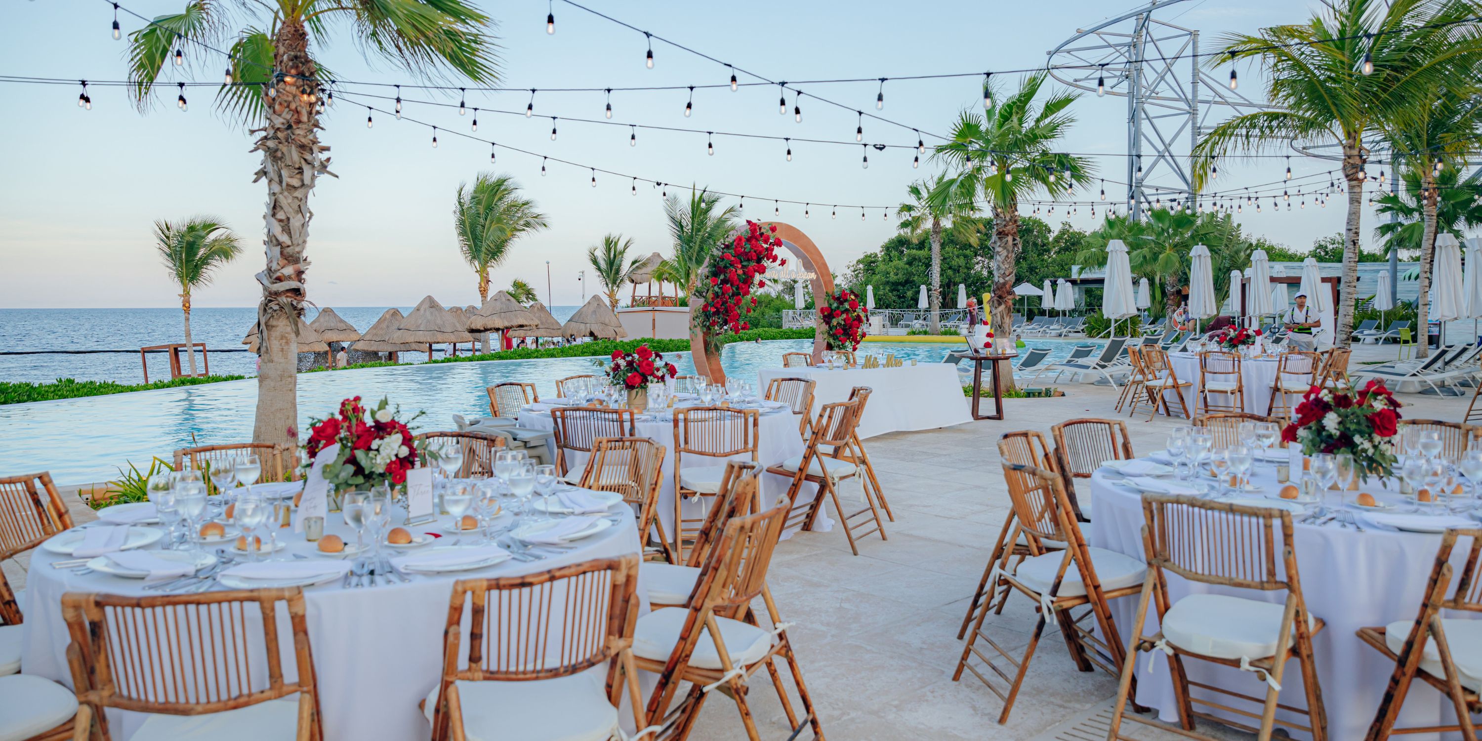 Destination wedding reception featuring round tables, floral centerpieces, a pool, and ocean backdrop.
