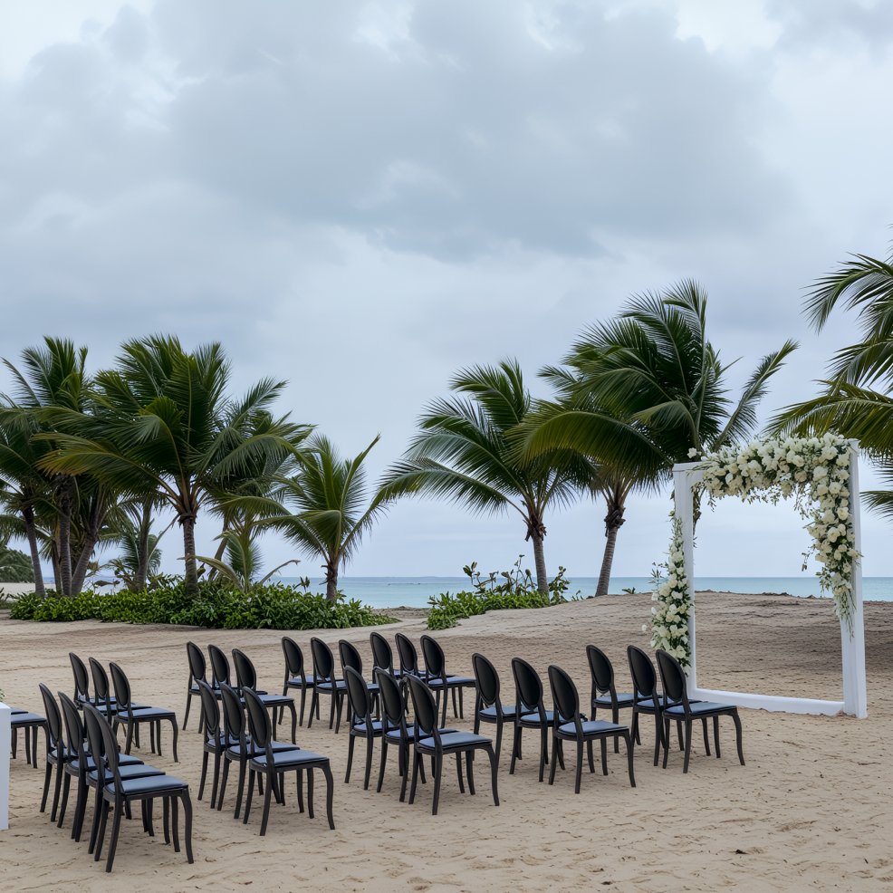 Black chairs are lined up for a destination wedding on a sandy beach with palm trees and a floral arch.