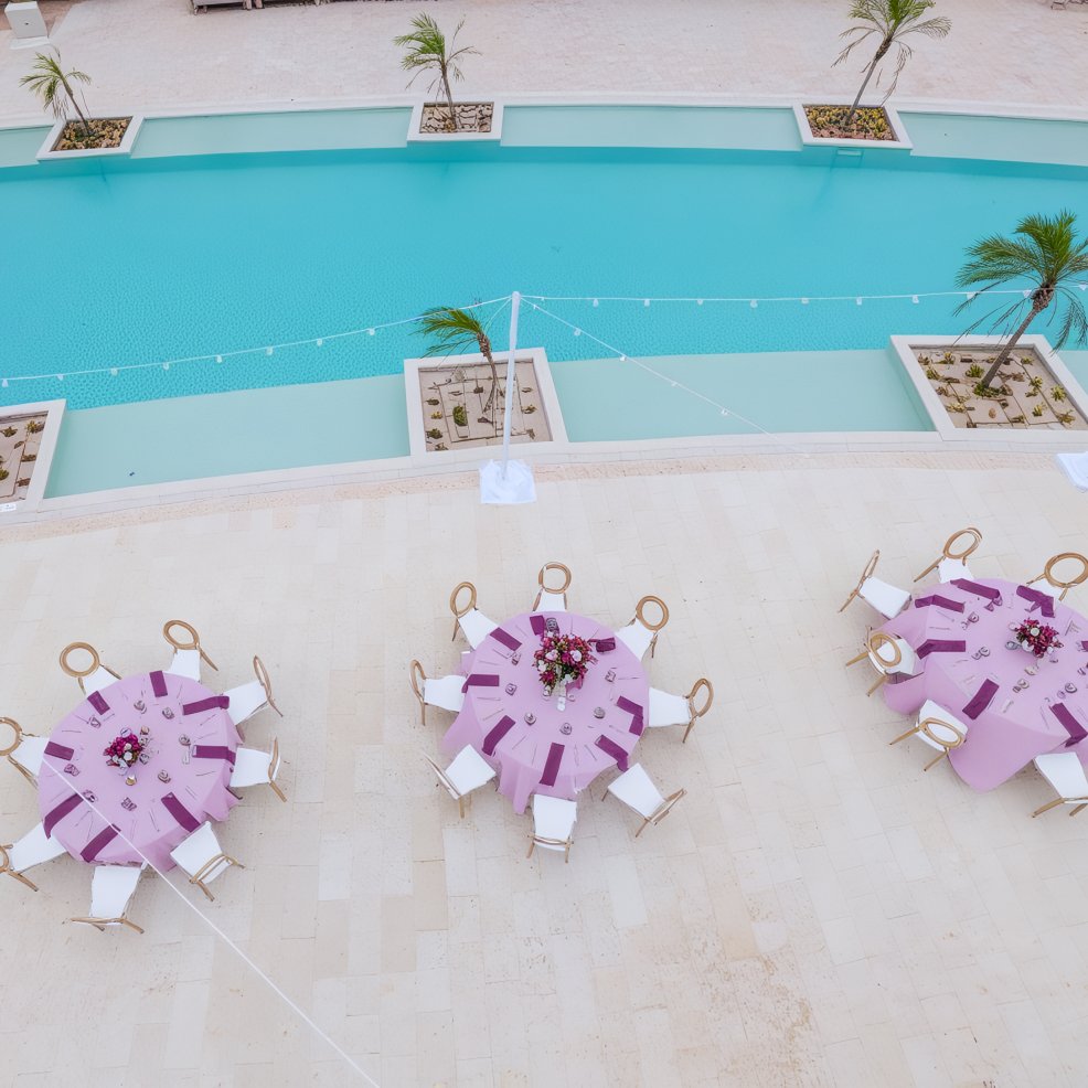 Three round tables with purple tablecloths set near a pool, viewed from above at a destination wedding.