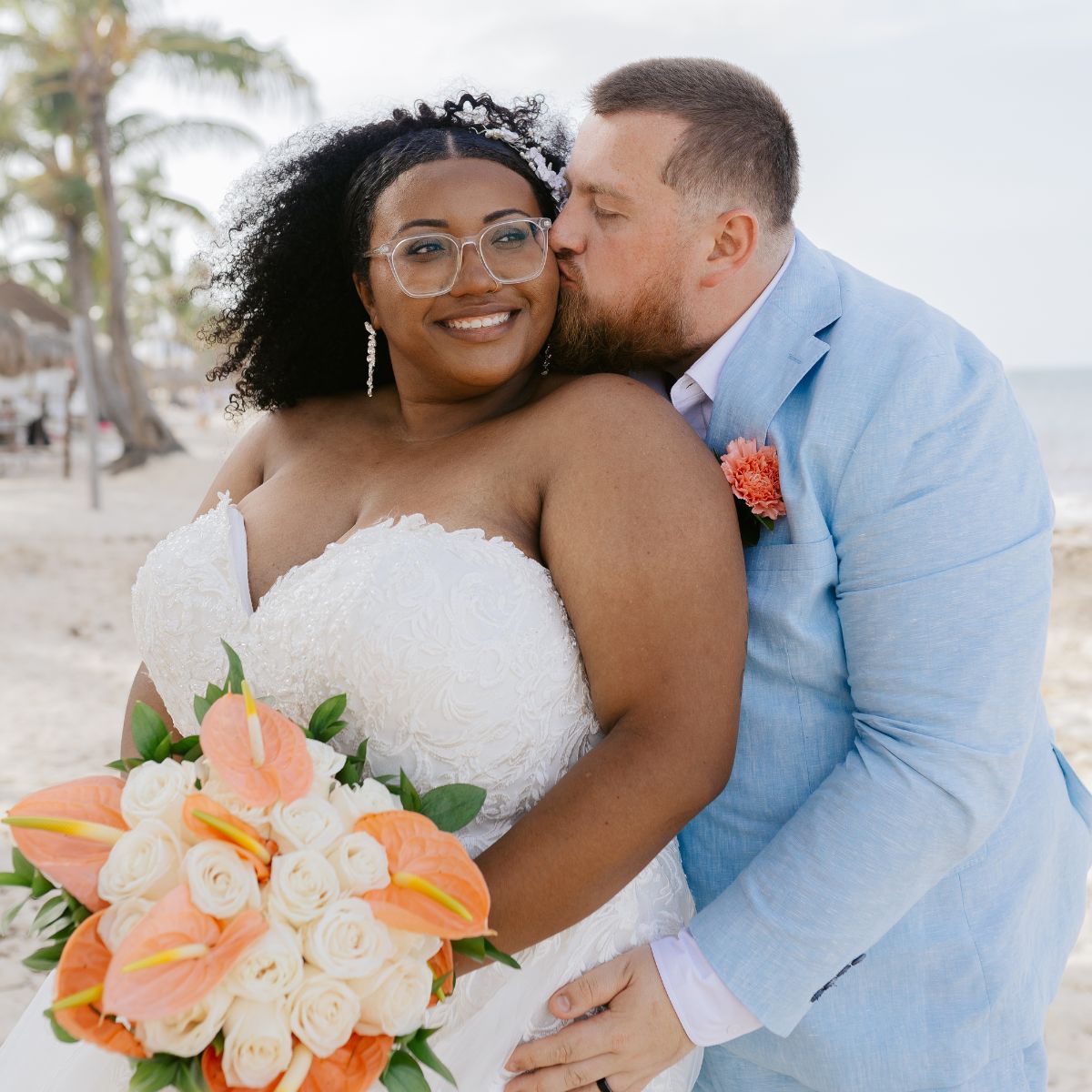 Alicia and Brian share a kiss on the cheek during their beach destination wedding; she holds a bouquet.