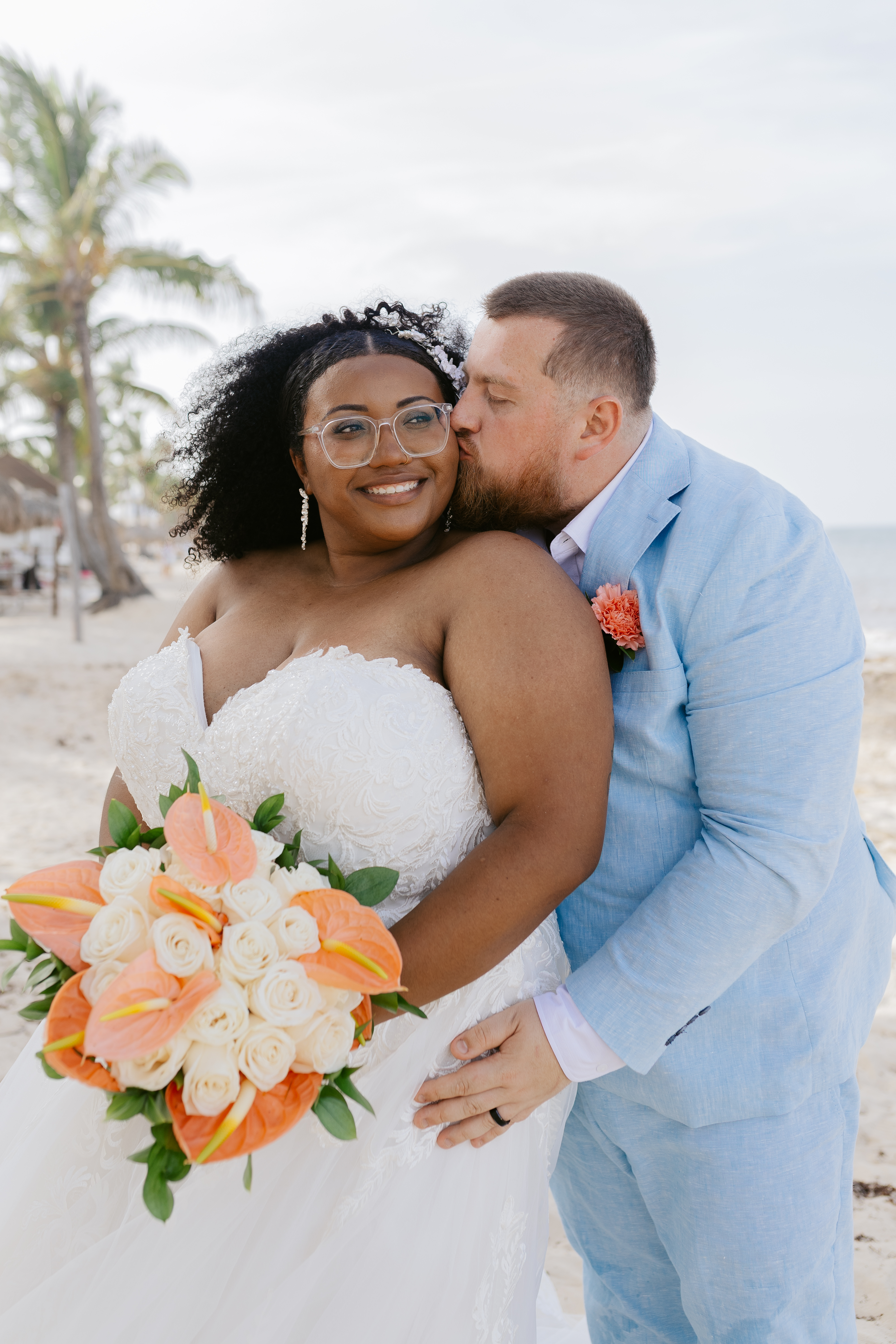 At a destination wedding on the beach, a bride with her bouquet smiles as the groom kisses her cheek.