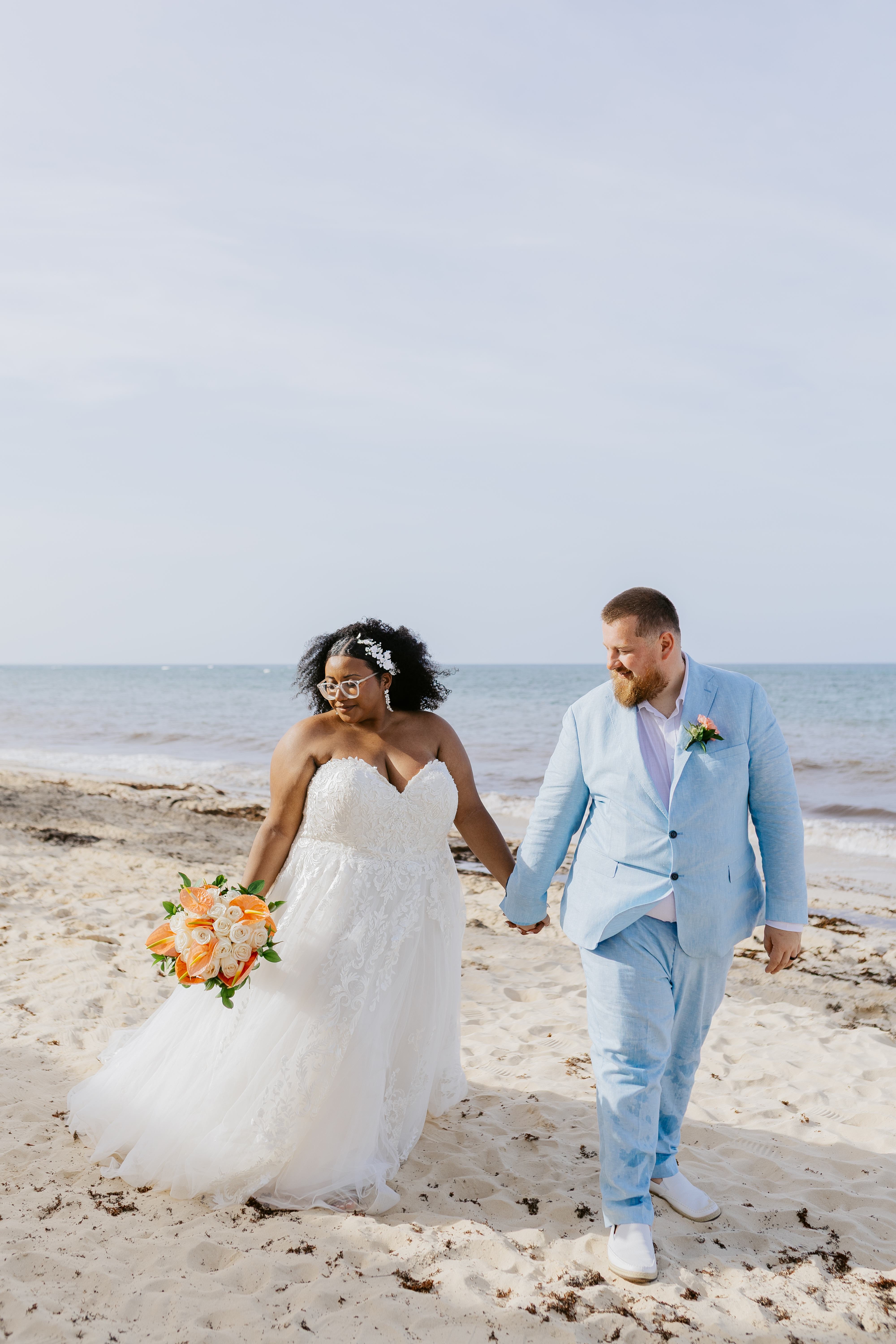 Alicia and Brian holding hands, walking on a sandy beach by the ocean at their destination wedding.