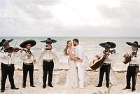 Teya and Charles embrace on a beach during a destination wedding as a mariachi band plays nearby.