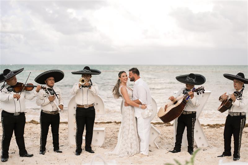 Teya and Charles embrace on a beach during a destination wedding as a mariachi band plays nearby.