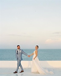Teya and Charles holding hands on a white terrace by the sea at their destination wedding under a clear sky.
